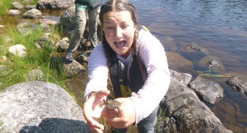 A person standing on the shore of a body of water holds a frog, who is trying to leap from their hands. 